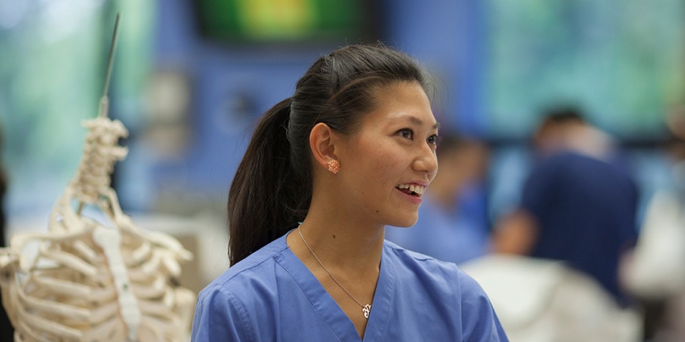 Student on scrubs in front of skeletal model.